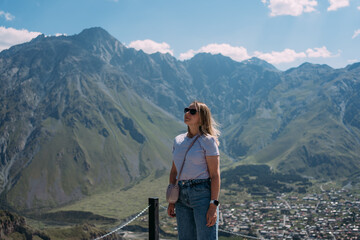 A tourist girl against the background of the mountains and the village of Stepantsminda in Georgia. The girl is wearing a purple T-shirt, denim shorts, sunglasses and a purple bag. Space, gorgeous vie