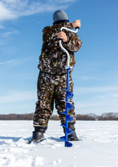 Naklejka premium A man in camouflage clothing is standing in the snow with a blue ice auger