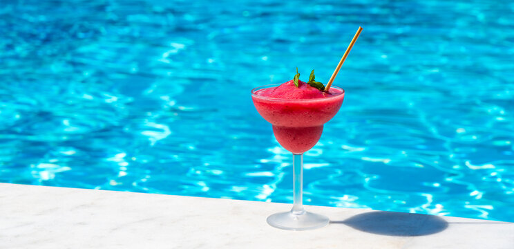 Red frozen drink in a cocktail glass placed on the edge of a bright blue swimming pool under the sun. Concept of cool tropical refreshment and relaxation in a summer setting