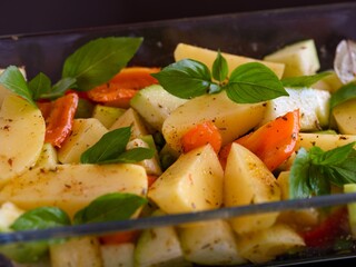 Vegetable pieces - potato, zucchini, pepper and green basil in a glass baking tray ready to cook. Close-up