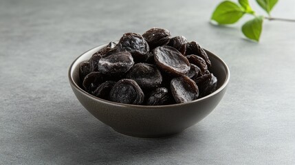 Obraz premium Dried salted plums in a black bowl on a gray surface with fresh greenery as a background. Ideal for culinary and food photography.