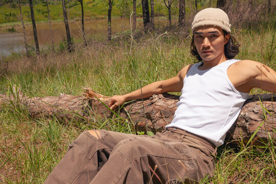An asian handsome man with long hair wearing white tank top, beige stylist hat reclines on a log, surrounded by a serene field under a clear sky, enjoying a moment of relaxation in nature. - Powered by Adobe