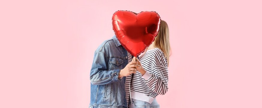 Happy young couple kissing behind heart-shaped balloon on pink background. Valentine's Day celebration