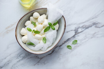 Bowl with mini mozzarella balls and green basil leaves, horizontal shot on a white stone background, high angle view