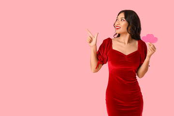 Beautiful young happy Asian woman with paper hearts pointing at something on pink background. Valentine's Day celebration