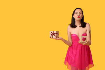 Beautiful young woman with gift boxes blowing kiss on yellow background. Valentine's Day celebration