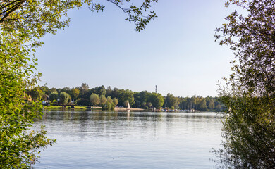 Obraz premium A serene lakeside view with a sandy beach, sailboats, and lush green trees under a clear sky, showcasing a peaceful outdoor recreation scene.