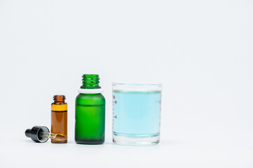 Three glass bottles in varying sizes, including a green bottle and a small amber vial, alongside a transparent glass of liquid, against a light background.