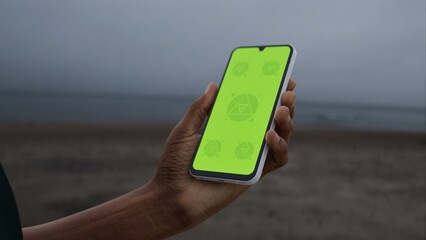 GREEN SCREEN CHROMA KEY Hand holding smartphone with green screen by the beach on an overcast day, perfect for customizable content placement in a coastal setting