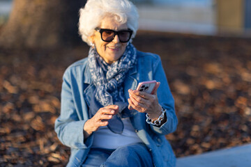white-haired elderly woman wearing denim sitting outside and looking at screen of mobile phone