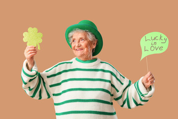 Happy senior woman in leprechaun hat holding decor for St. Patrick's Day celebration on beige...