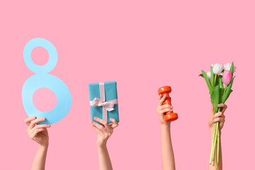 Female hands with paper figure 8, dumbbell, gift box and tulip flowers for International Women's Day on pink background