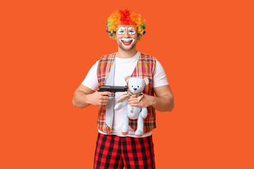 Young man as crazy clown with gun and toy bear on orange background
