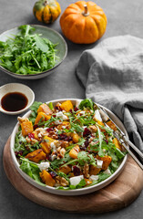 Autumn salad with pumpkin, arugula, feta cheese, nuts and dried berries in a plate on wooden board on dark background