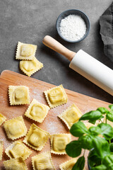 Homemade pasta ravioli with filling on wooden cutting board on dark background with flour, basil and rolling pin.
