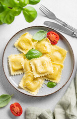 Flat lay of homemade ravioli pasta with cheese, tomatoes and basil in a plate on a light  background with  fork, herb and napkin.