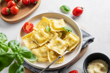 Ravioli pasta with parmesan, tomatoes and basil in a plate on a napkin and wooden board on a light  background