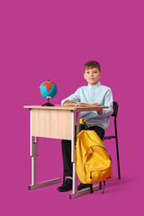 Cute boy sitting at school desk on pink background