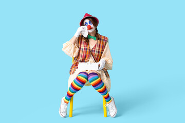 Young woman dressed as sad clown crying on chair against blue background. April Fool's day celebration