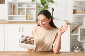 Happy young woman holding box with chocolate covered strawberries in kitchen