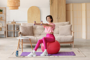 Beautiful young woman training with dumbbells on fitball in living room