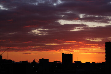 The predawn sky is illuminated by a brilliant orange background, creating fiery hues of blood red orange in the lingering morning clouds as a silhouetted city silently slumbers below.