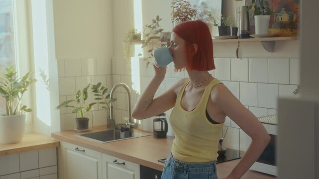 Young woman with bright red hair standing in cozy kitchen basking in sunlight, drinking coffee from cup and looking pensively through window in morning at home