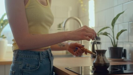Young beautiful woman with red hair stirring coffee in cezve as cooking drink on stovetop in kitchen filled with sunlight during morning at home. Tilt-up, close-up shot