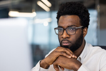 Obraz premium African American man businessman intensely focused on computer in office. Wearing glasses, displaying thoughtful expression. Represents concentration, work, technology.