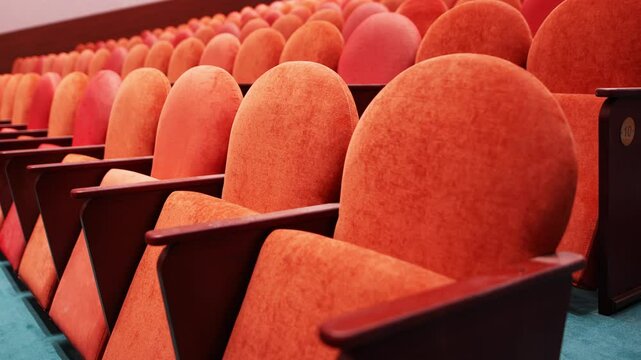 Rows of red colored velvet seats arranged from bottom to top in hall. Wooden armchairs stand near wall decorated with large and rich Tatar patterns