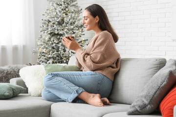 Beautiful young woman with glass cup of hot cocoa sitting on sofa in living room on Christmas eve