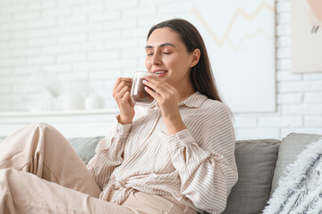 Beautiful young woman with glass cup of hot cocoa sitting on sofa in living room