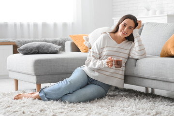Beautiful young woman with glass cup of hot cocoa sitting on carpet in living room