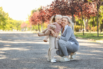 Cute little girl with toy bear and her mother hugging in park on autumn day