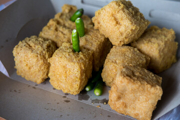 Close-up of crispy, golden-brown fried tofu in a cardboard container with green chili peppers. The texture and savory appeal are highlighted.