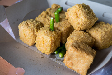 Close-up of crispy, golden-brown fried tofu in a cardboard container with green chili peppers. The texture and savory appeal are highlighted.