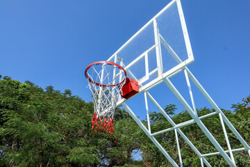 basketball hoop with a red rim and white net, mounted on a glass backboard, set against a blue sky and green trees. low angle view