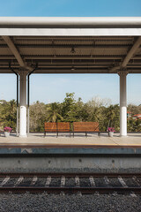 Fototapeta premium Quiet tropical train station platform with wooden benches, potted flowers, and lush palm trees in the background. A serene setting perfect for travel, transportation, and scenic getaway themes.