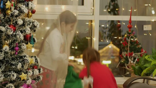 Small children in Christmas pajamas are holding green socks for gifts in their hands, waving them at each other, fighting, laughing, having fun near the window and the Christmas tree. Lots of lights