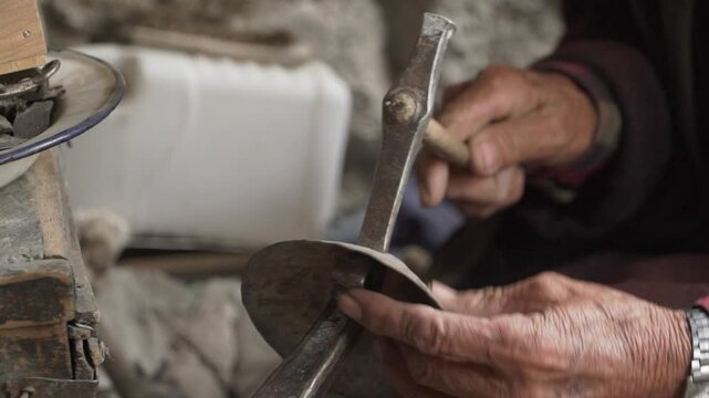 An elderly blacksmith meticulously crafts copper using a hammer and anvil in a dimly lit workshop. Ladakh, India