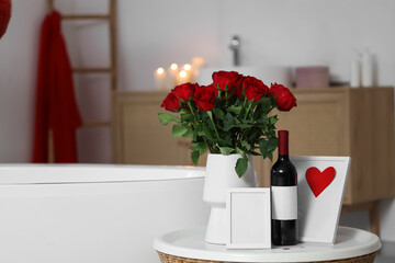 Vase with roses, wine and blank frame on table in bathroom decorated for Valentine's Day