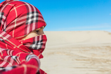Portrait of a young girl wearing a red shawl covering her face against a background of blue sky and sand. Concept of hope and emigration in the Middle East.