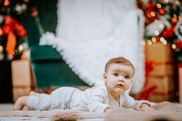  Baby lying on a soft blanket, looking upward, surrounded by festive Christmas decor, twinkling lights, and wrapped presents in a cozy home.