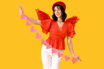 Beautiful young happy woman dressed as cupid with heart-shaped garland on yellow background. Valentine's Day celebration