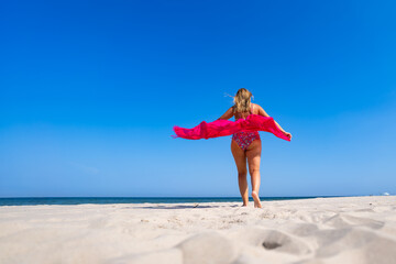 Beautiful middle aged woman in bathing suit walking with scarf fluttering in wind on sandy beach in summer. Back view