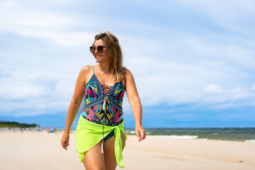 Beautiful middle-aged woman in swimsuit walking on sandy beach in summertime. Front view