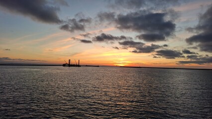 Sunset view over a calm sea with silhouettes of industrial structures and ships on the horizon. Dramatic clouds and vibrant orange and blue hues create a serene yet dynamic scene