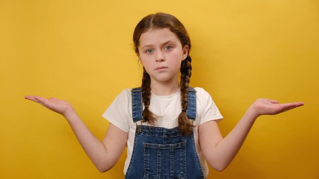 Portrait of confused preteen girl child show spread hands oops gesture, posing isolated over plain yellow color background wall in studio. Childhood lifestyle concept