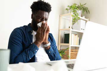 Sick Black man working from home office. Male blowing nose with tissue while working with laptop.