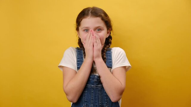 Portrait of shocked preteen caucasian girl child cover palms mouth oops gesture, posing isolated over plain yellow color background wall in studio. Childhood lifestyle and emotion concept